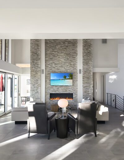 Modern living room interior with a stone fireplace and mounted television, featuring minimalist furniture and abundant natural light.
